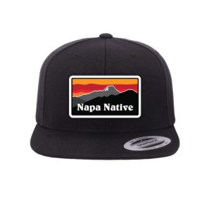 The Grape Crusher Snap Back is a black hat with a snapback closure, featuring a rectangular patch of a mountain sunset and bold white "Napa Native" lettering.